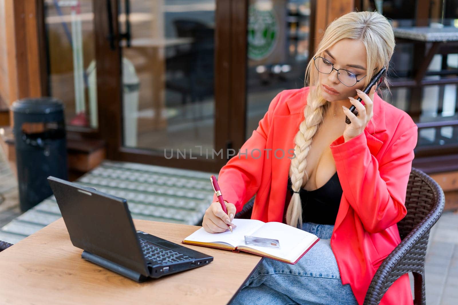 Beautiful woman sits at table in street cafe and works on tablet. Concept remote work, free work schedule. In a bright pink jacket and glasses