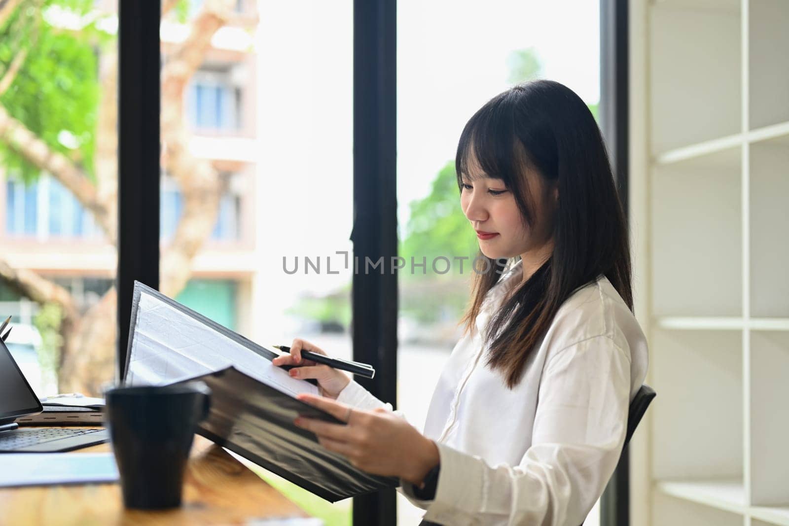 Asian female financier reading paper document, analyzing marketing data at desk in bright office.