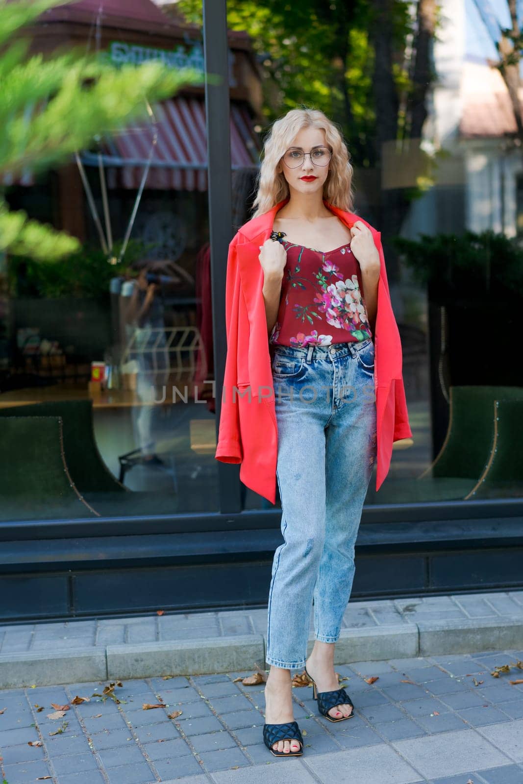 beautiful blonde woman in glasses dressed in red jacket and blue jeans posing on street in the city by EkaterinaPereslavtseva