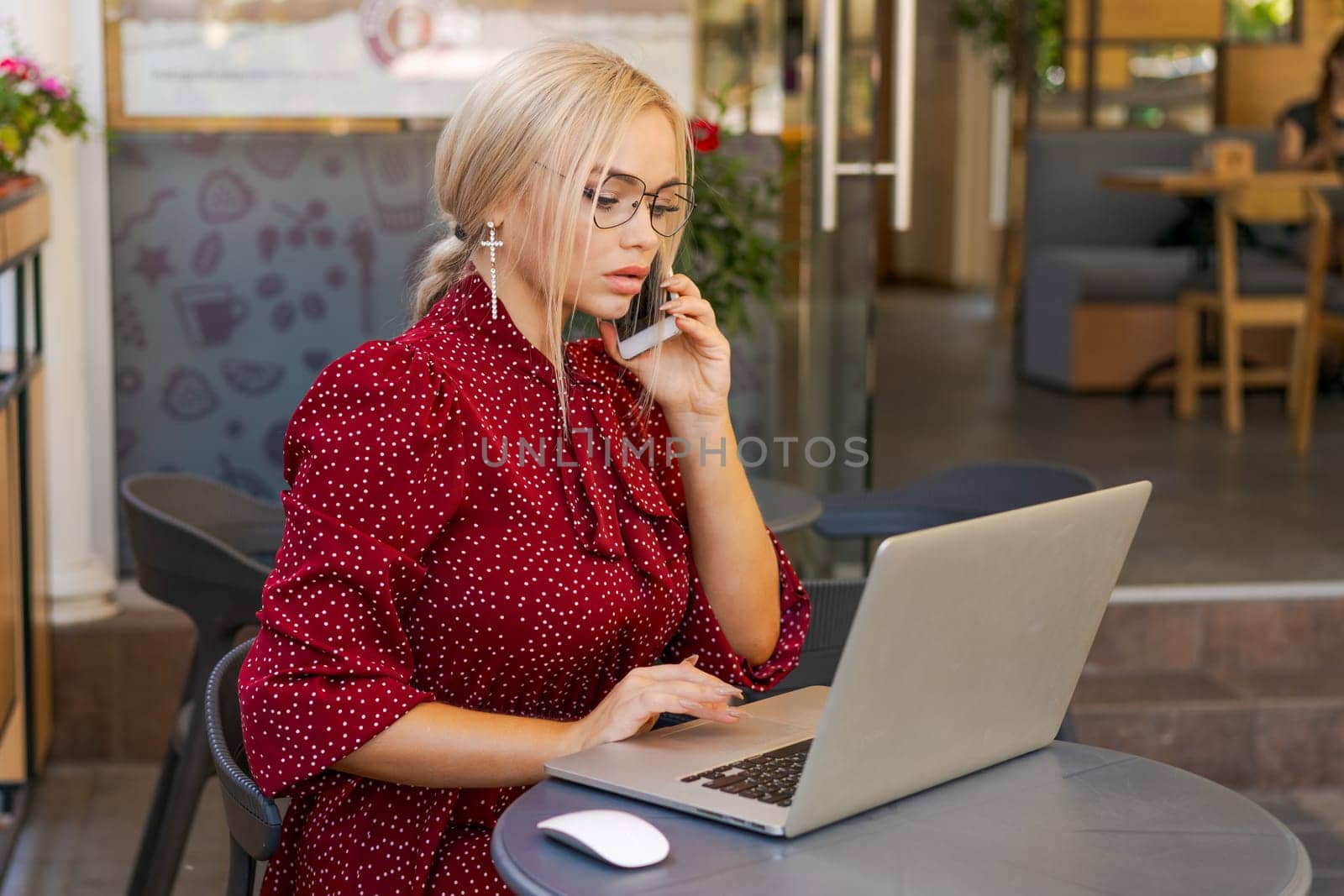 Beautiful woman works on laptop in coffee shop and using mobile phone in a red dress and glasses, business blonde online business