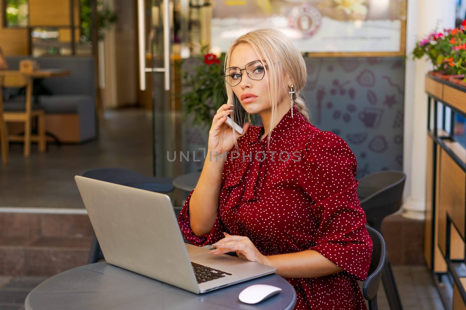 Beautiful woman works on laptop in coffee shop and using mobile phone in a red dress and glasses, business blonde online business