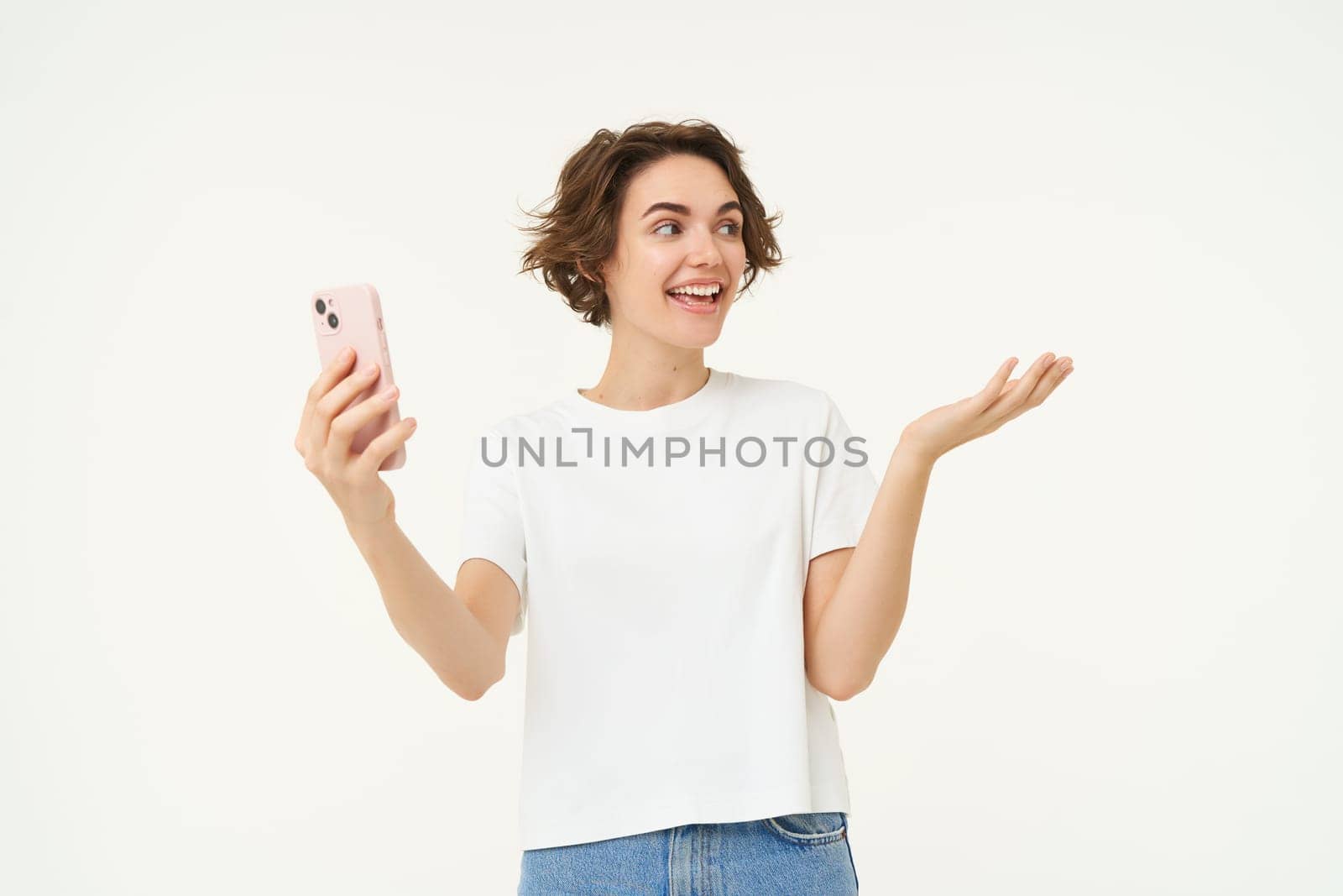 Potrait of woman video chats with friend, showing around, demonstrating something, has an online meeting on smartphone, standing over white background by Benzoix