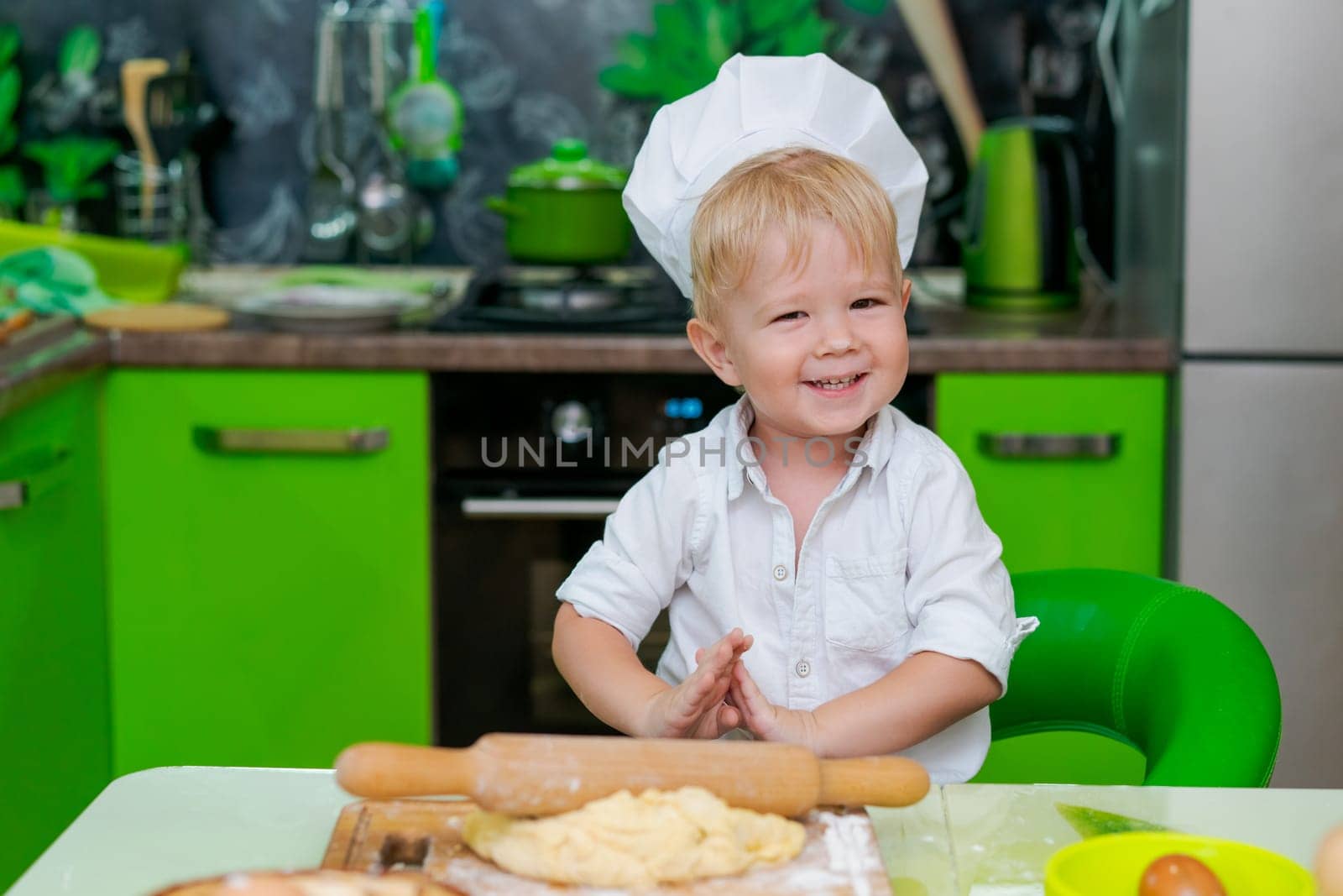 happy little boy preparing dough in kitchen at table. there are dough products on table, dressed as chef by EkaterinaPereslavtseva