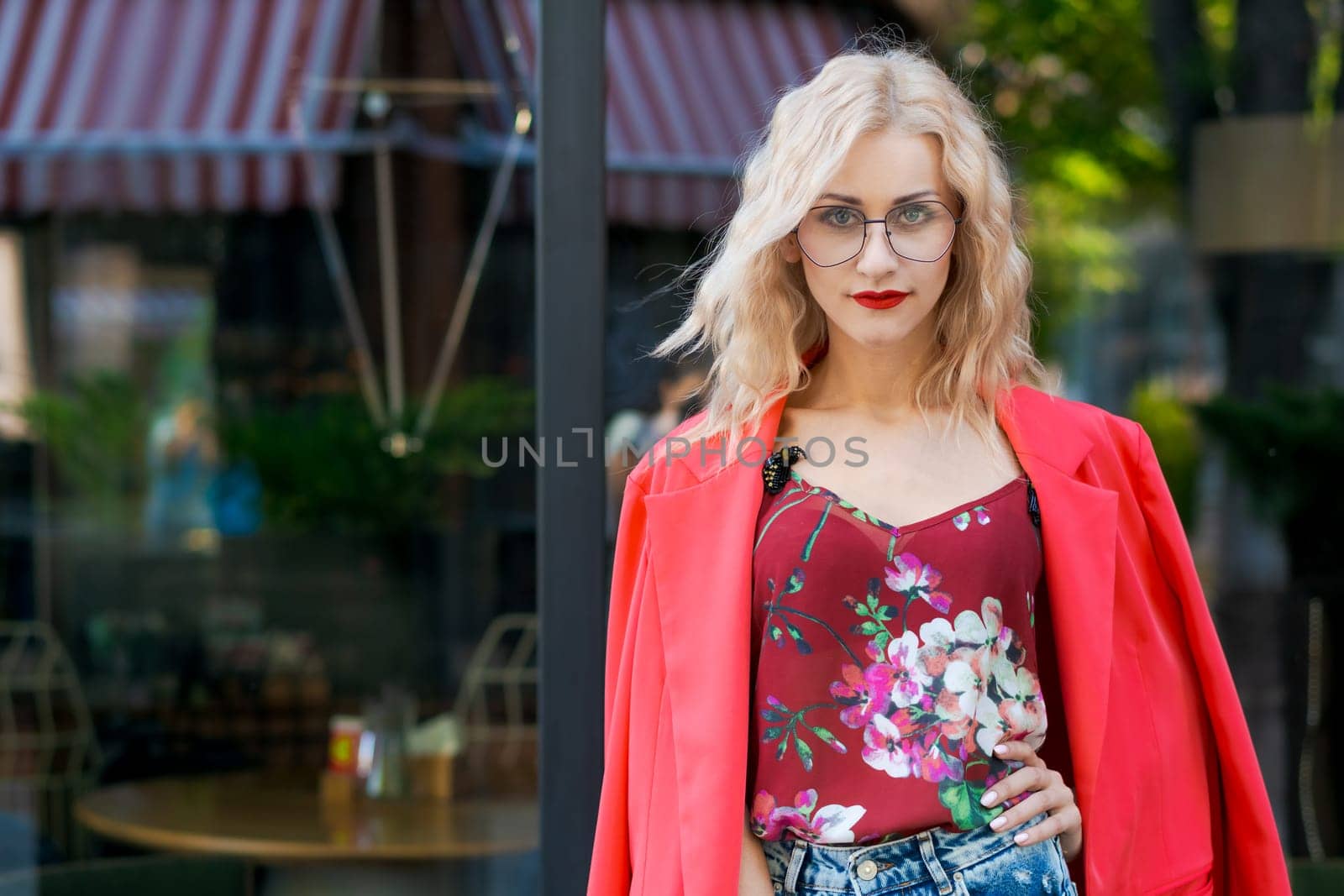 beautiful blonde woman in glasses dressed in red jacket and blue jeans posing on street in the city by EkaterinaPereslavtseva