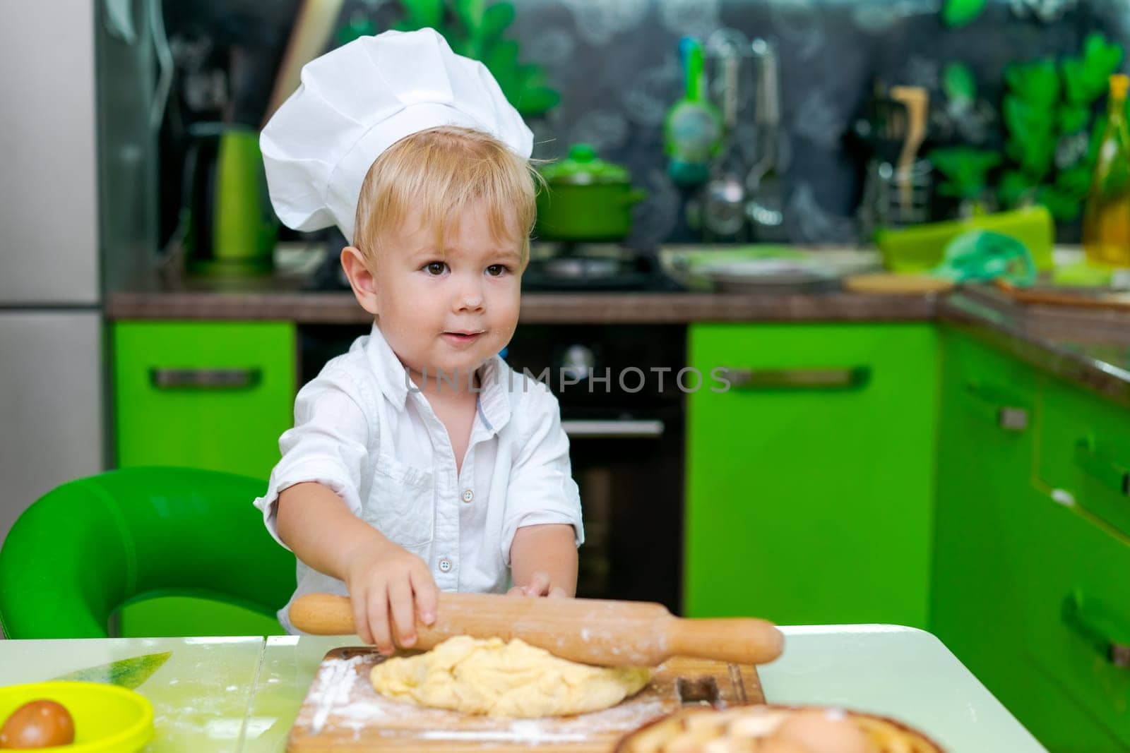 happy little boy preparing dough in kitchen at table. there are dough products on table, dressed as chef by EkaterinaPereslavtseva