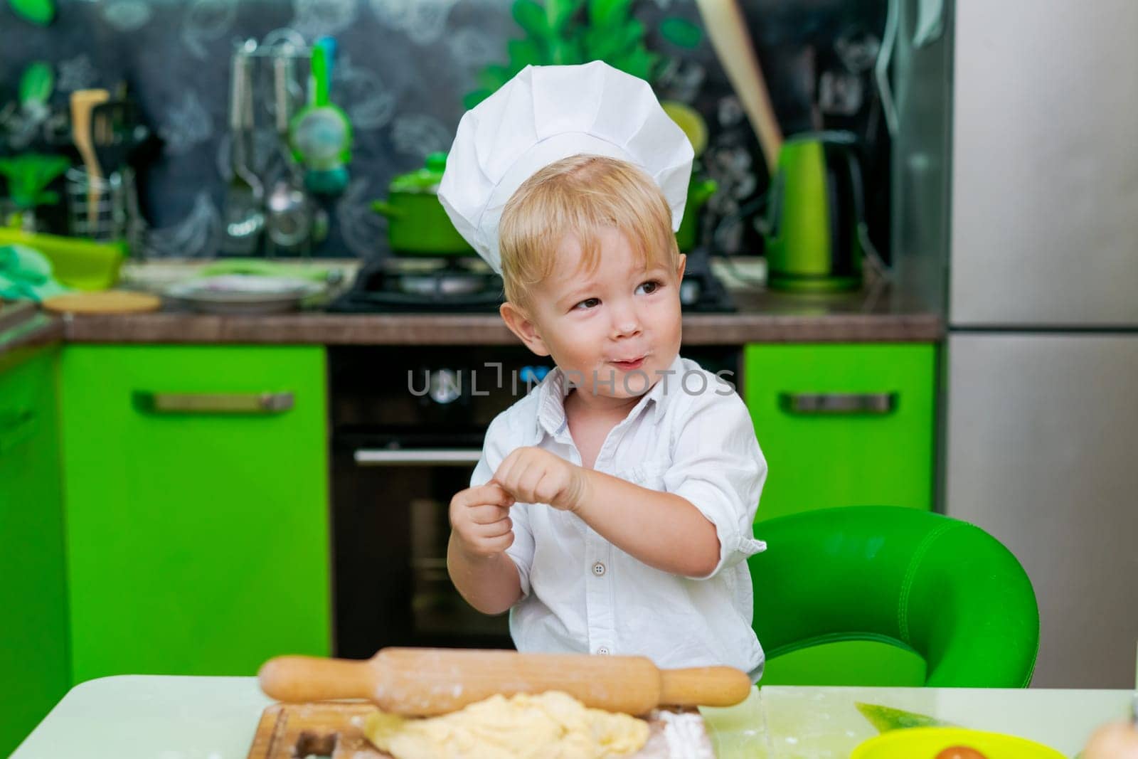 happy little boy preparing dough in kitchen at table. there are dough products on table, dressed as chef