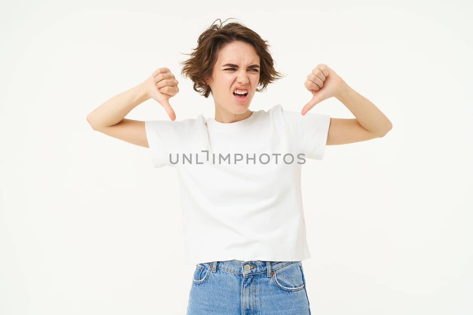 Portrait of disappointed young woman, student shows thumbs down from dislike, disapprove something bad, standing over white background.