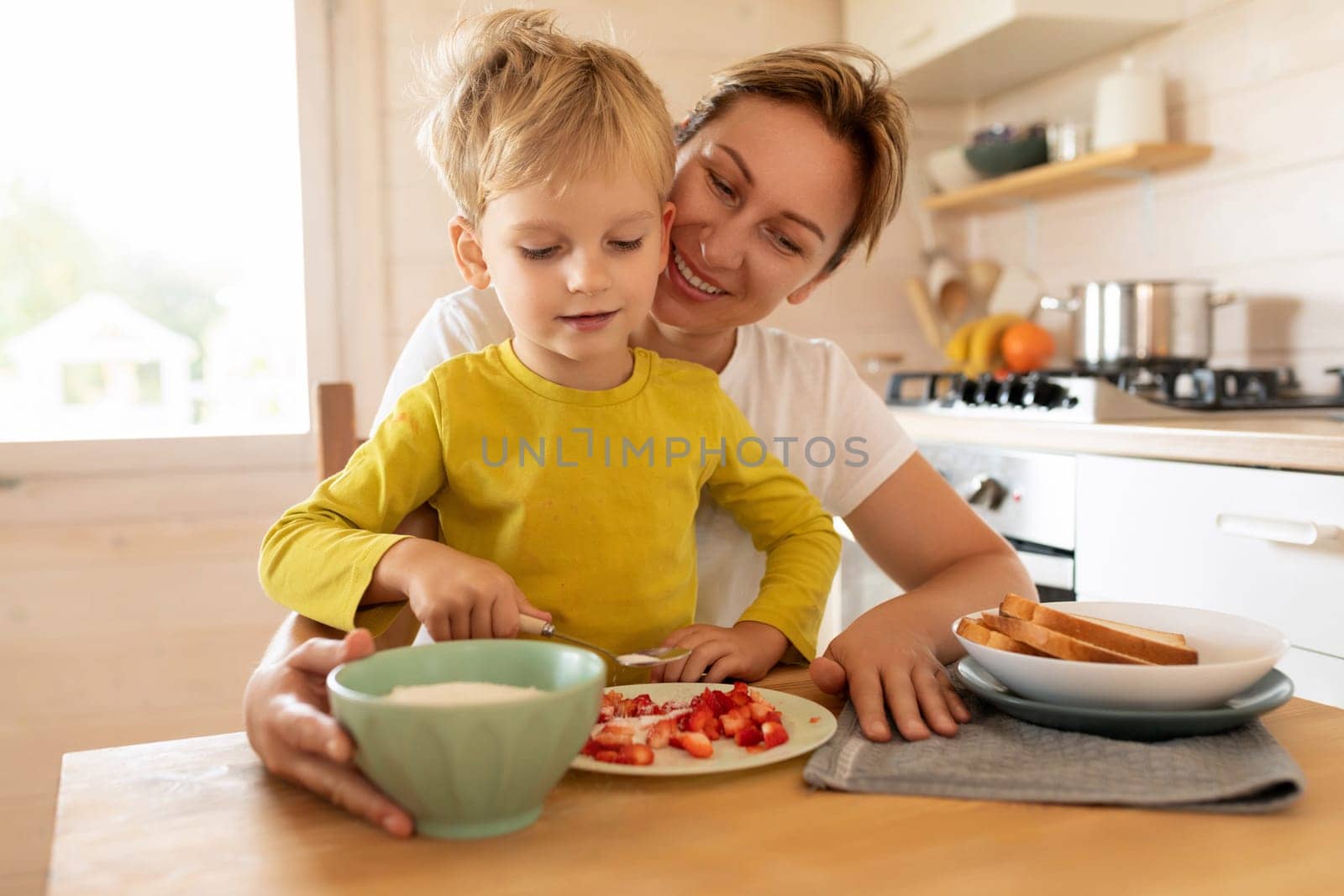 young caucasian blond mother and child son sitting in the morning in the kitchen and preparing breakfast with toast and strawberries by TRMK