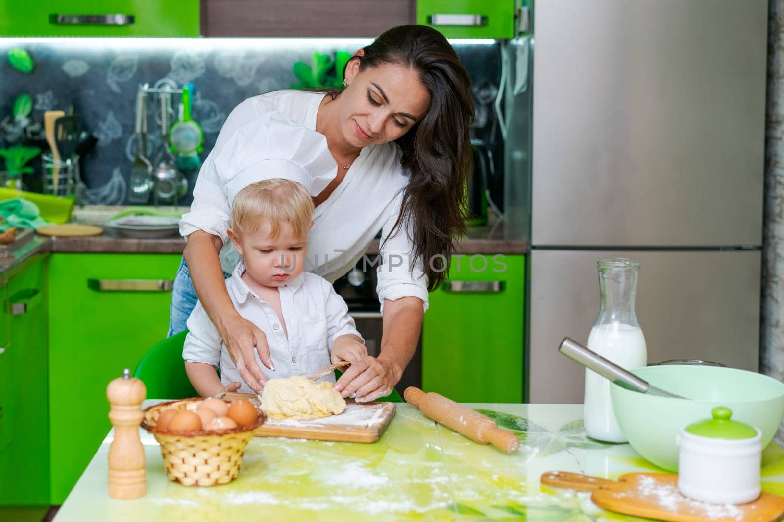 happy family mother and little son preparing dough in kitchen at table. products for dough are on table