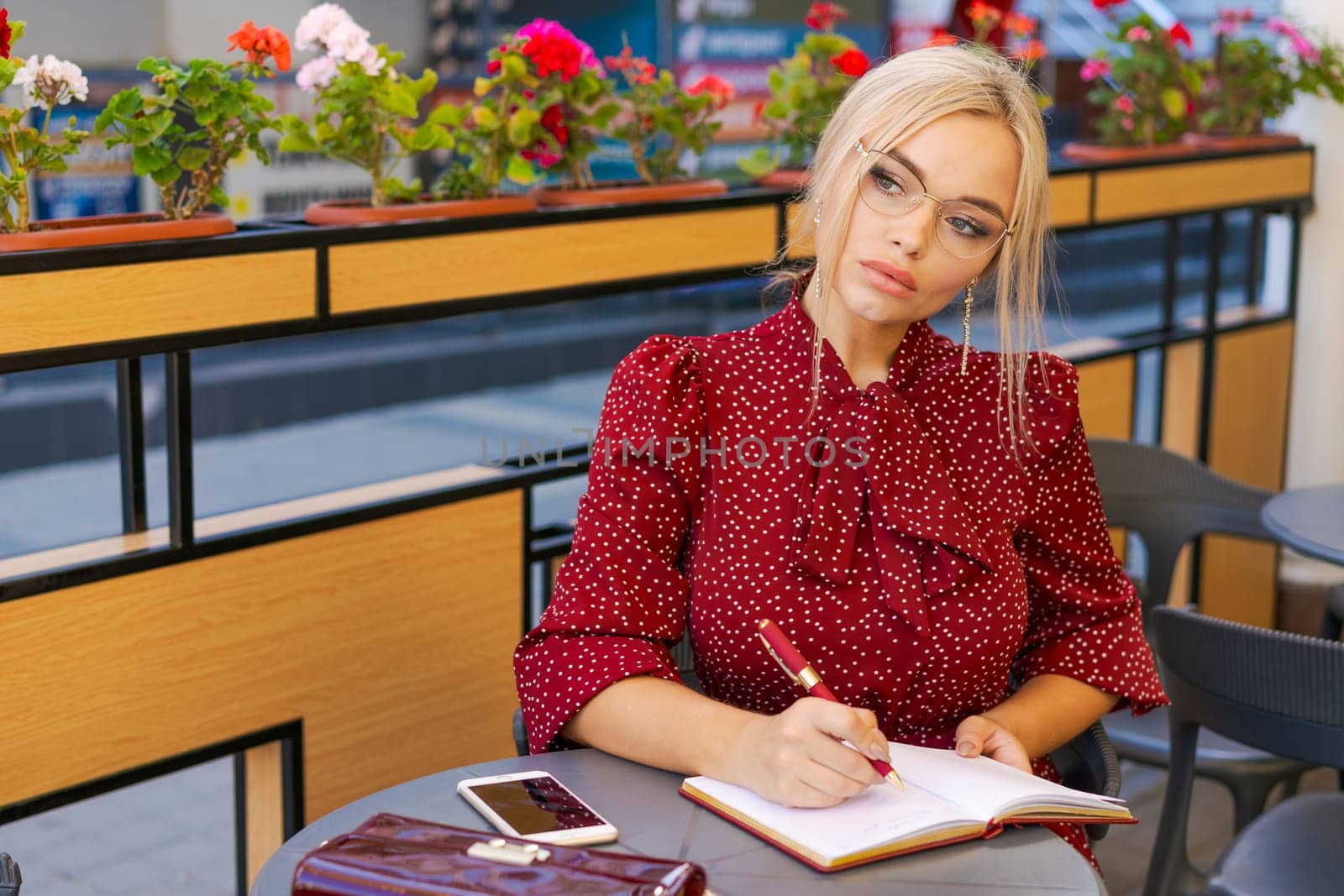 Nice caucasian woman writes in notebook spending time outdoors in cafe. Blonde girl wears a red dress. Inspiration concept and working with studies remotely