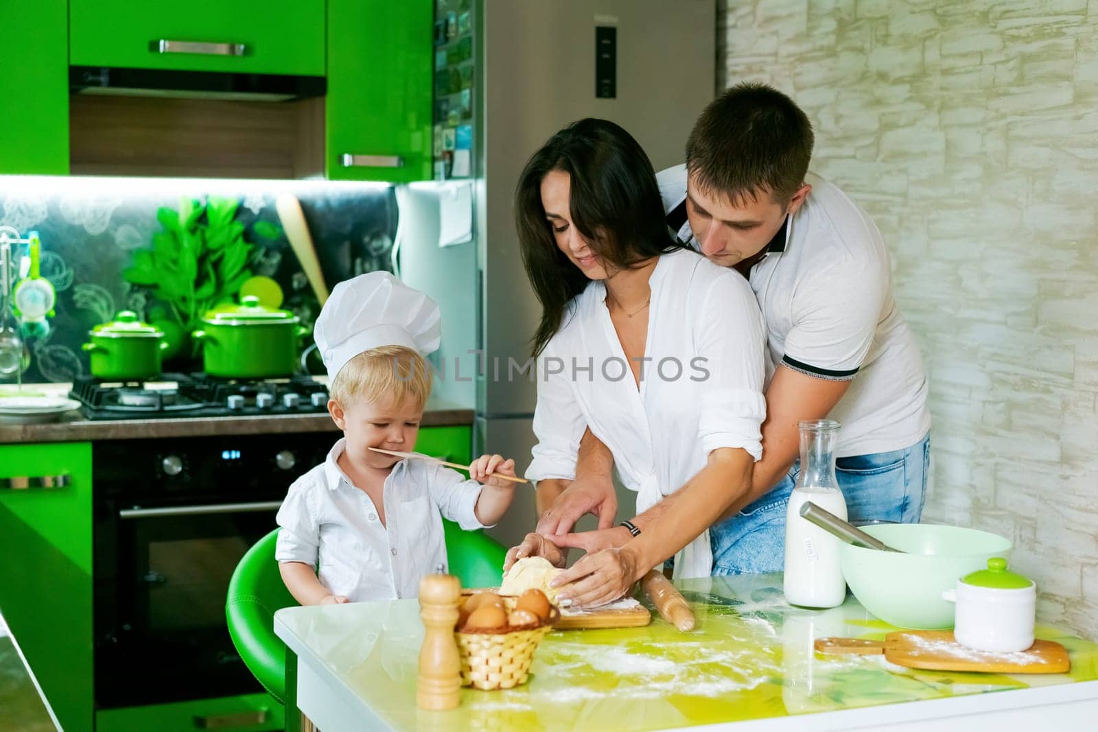 happy family mom and little son and dad are preparing dough in kitchen at table. products for dough are on table