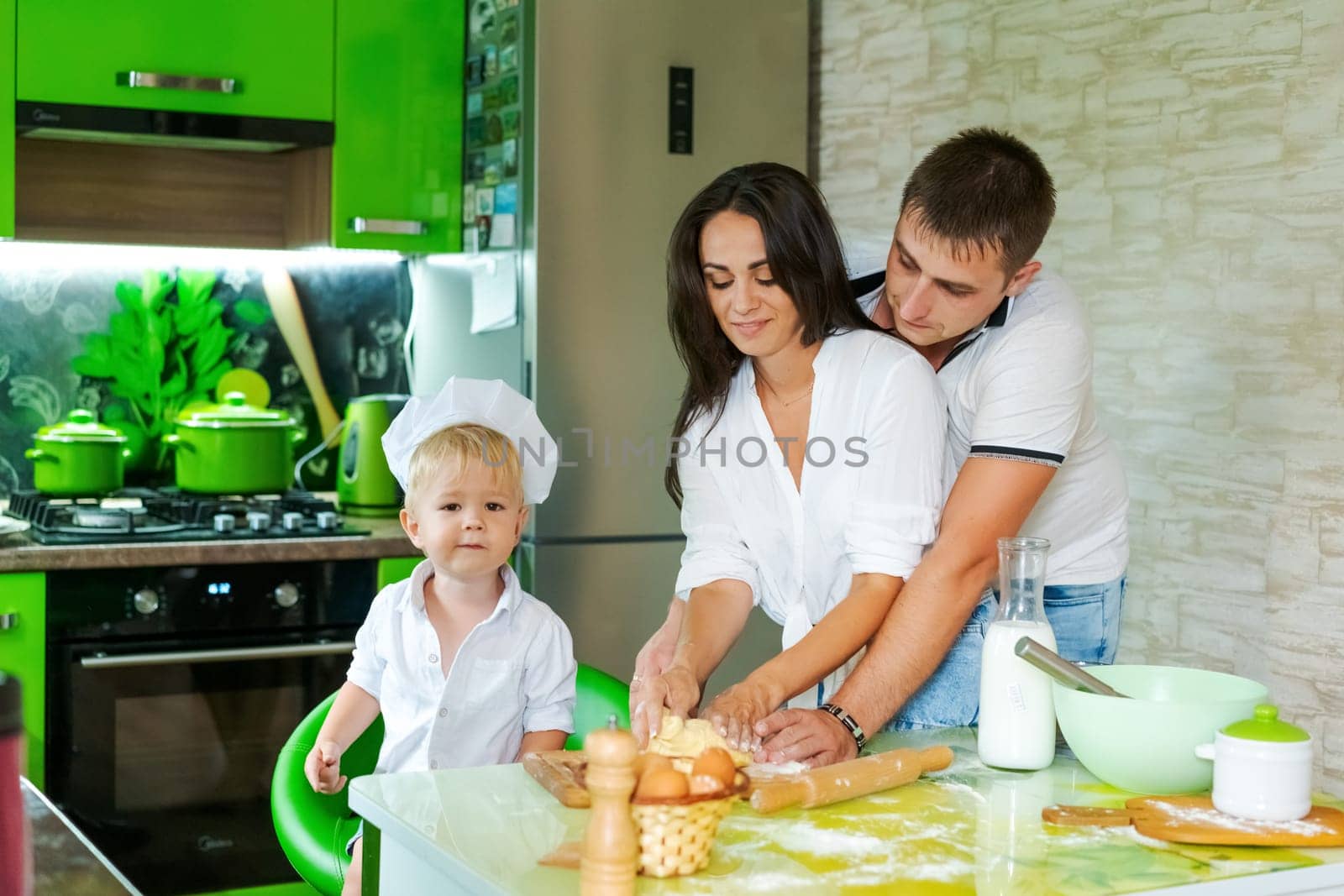 happy family mom and little son and dad are preparing dough in kitchen at table. products for dough are on table
