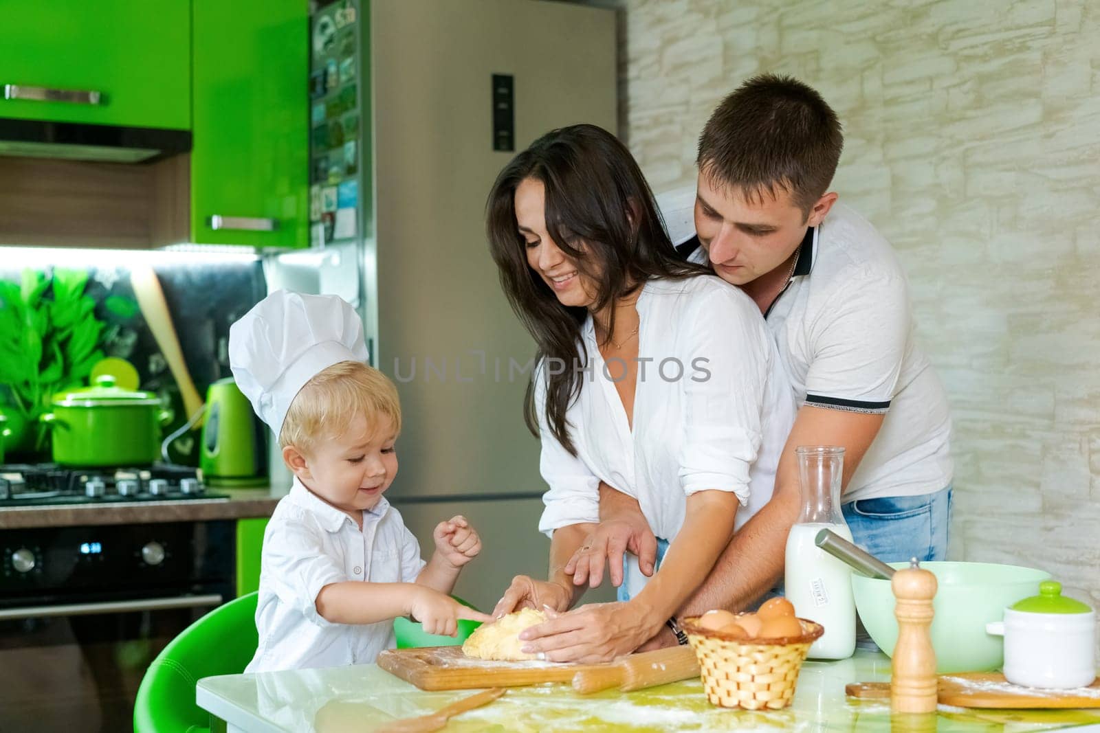 happy family mom and little son and dad are preparing dough in kitchen at table. products for dough are on table