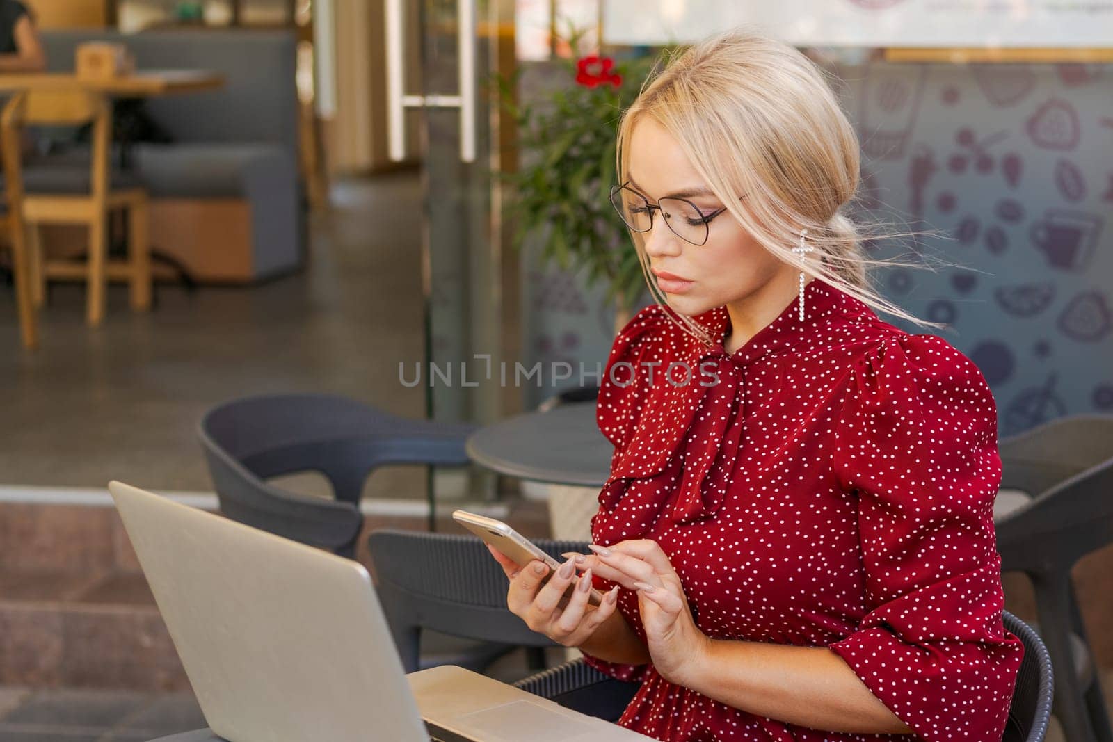 Beautiful woman works on laptop in coffee shop and using mobile phone in a red dress and glasses, business blonde online business