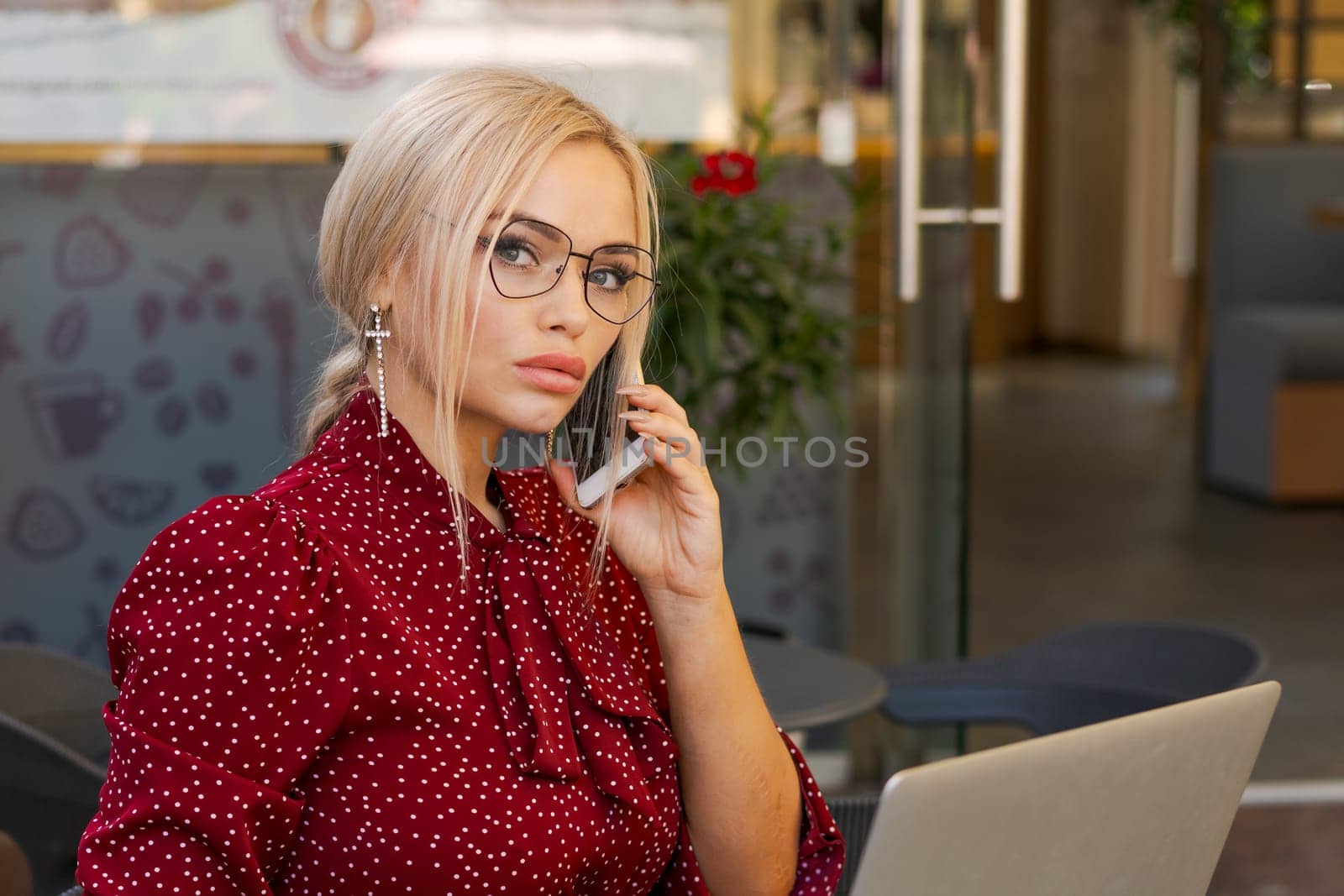 Beautiful woman works on laptop in coffee shop and using mobile phone in a red dress and glasses, business blonde online business