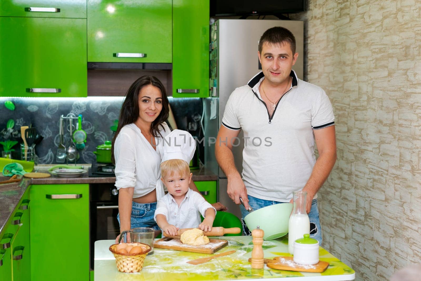 happy family mom and little son and dad are preparing dough in kitchen at table. products for dough are on table
