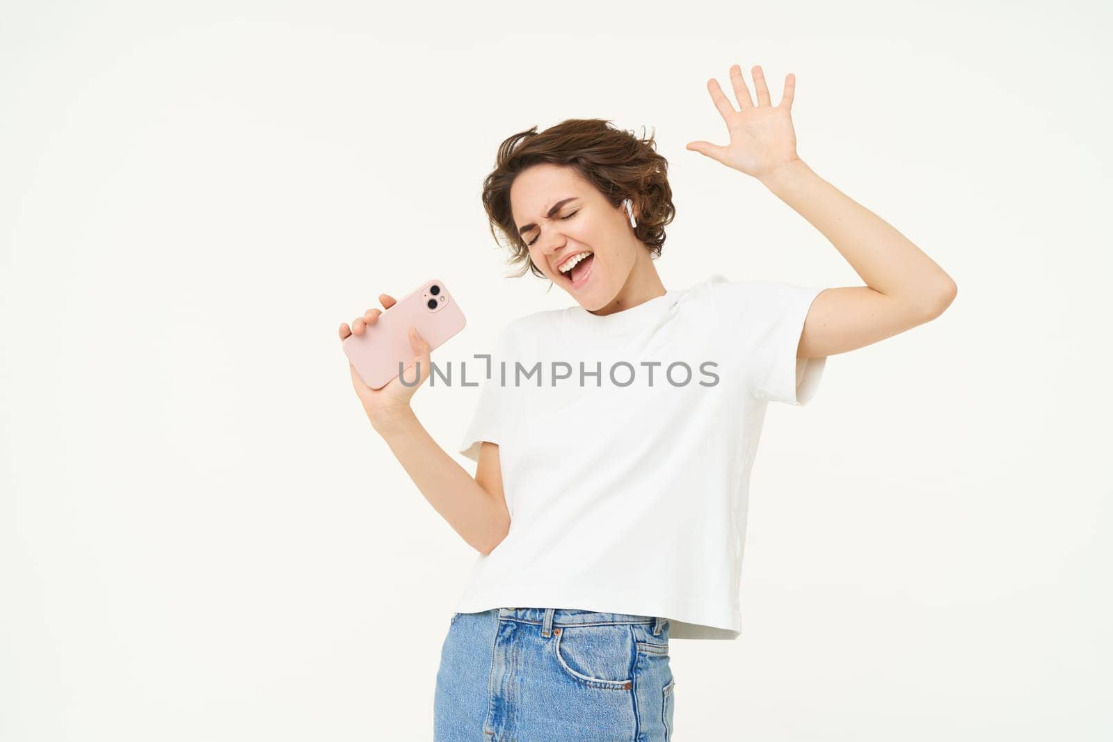 Portrait of carefree girl singing in wireless headphones into smartphone microphone, playing music game on mobile app, standing over white background.