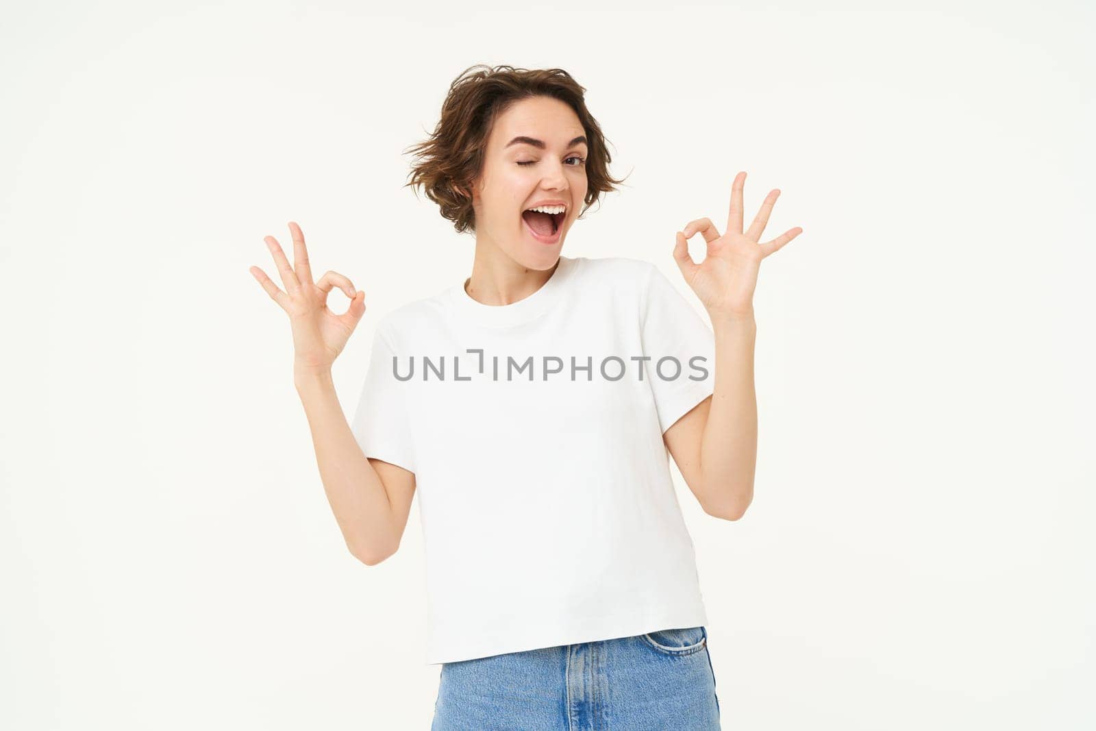 Portrait of young woman, showing okay, ok gestures and smiling, giving positive feedback, recommending, standing over white background.