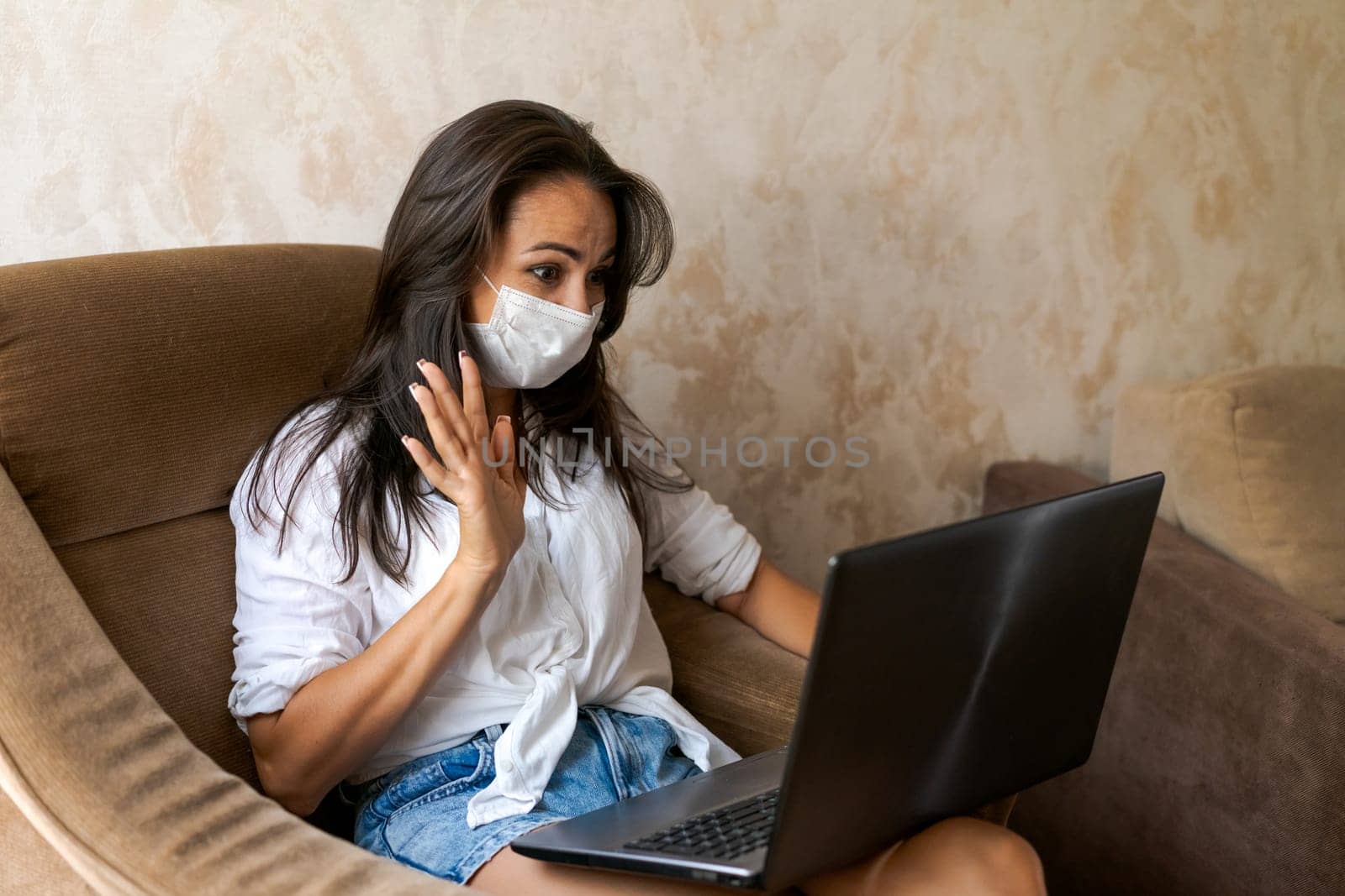 Cute woman at home in chair with laptop wearing protective mask on her face communicates on an online conference, concept of working from home, freelancer or study