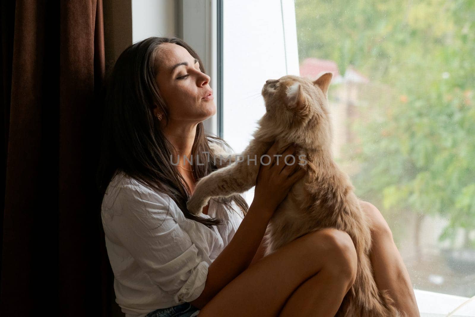 cute woman sitting on the windowsill with a red cat and looking out the window