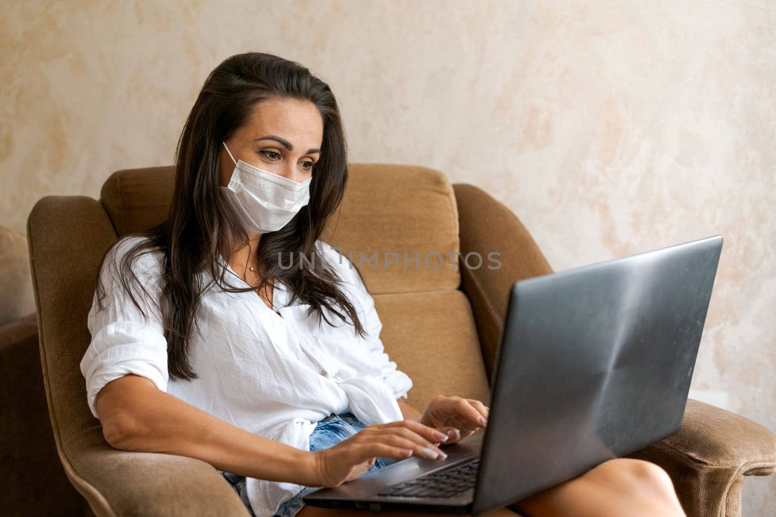 Cute woman at home in chair with laptop wearing protective mask on her face communicates on an online conference, concept of working from home, freelancer or study