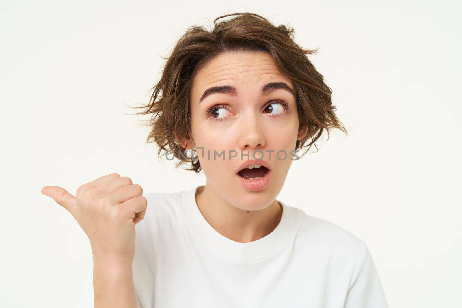 Image of girl with surprised face, points finger left, looks shocked, poses over white studio background. copy space