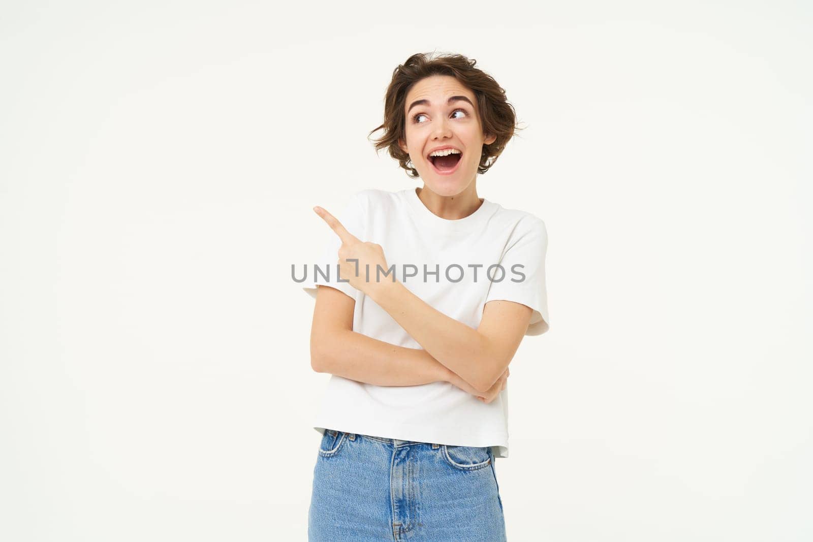 Excited brunette woman laughing and smiling, pointing finger left, showing advertisement, standing over white background.