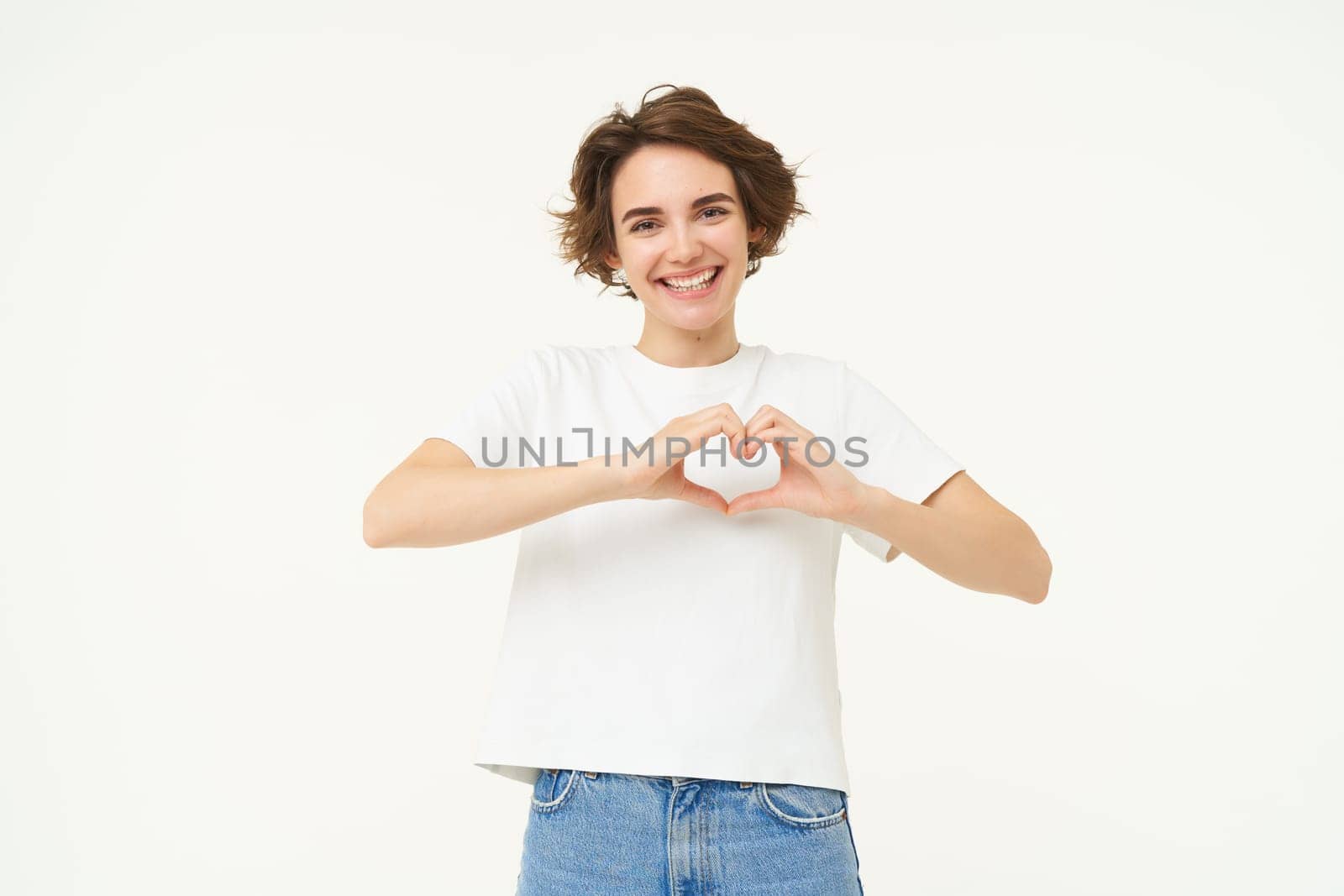 Portrait of young woman shows heart, love and care gesture, express sympathy, like something or someone, smiling happily, standing over white background.