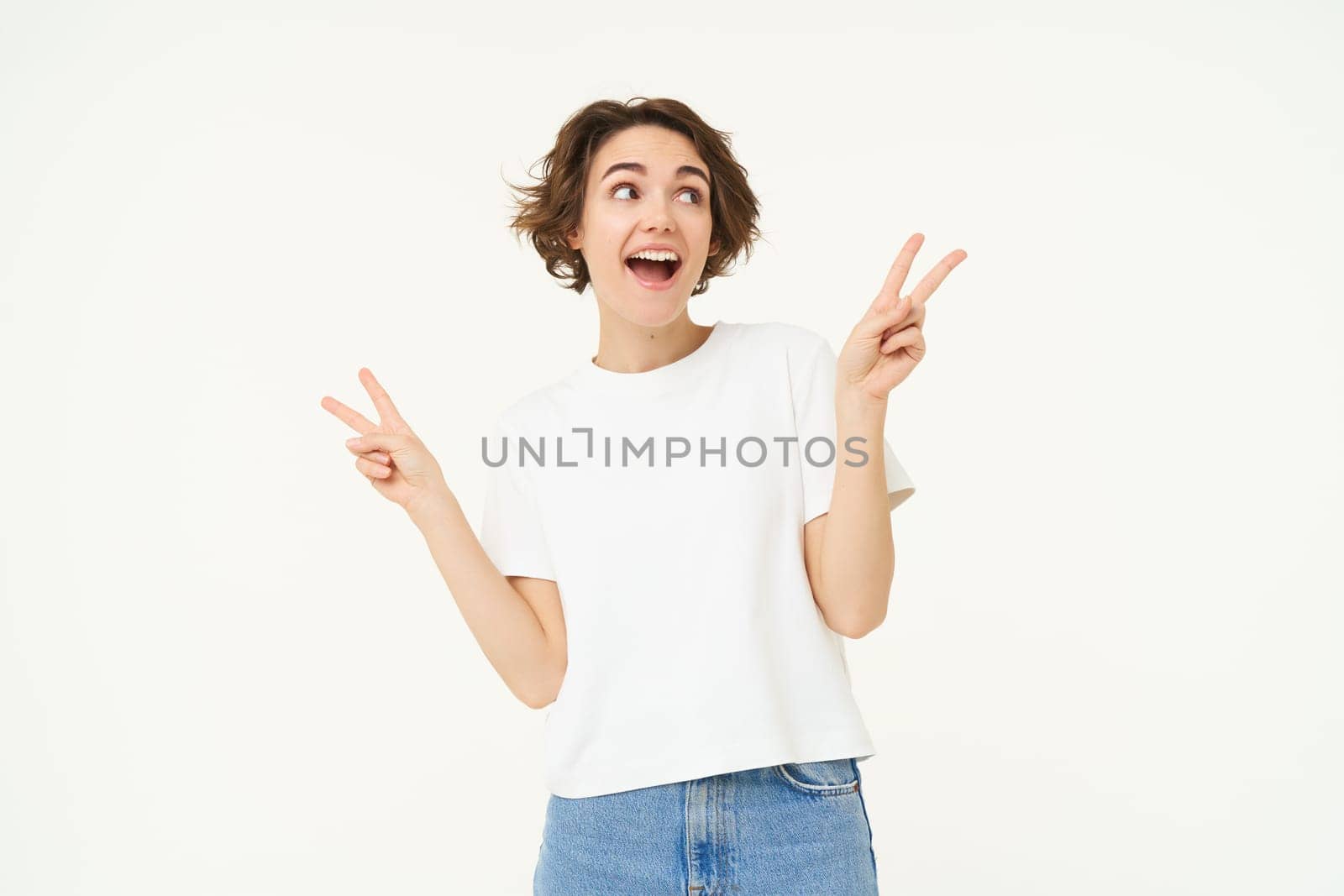 Portrait of young girl shows peace, v-sign and smiling, express posivity and joy, posing over white studio background.