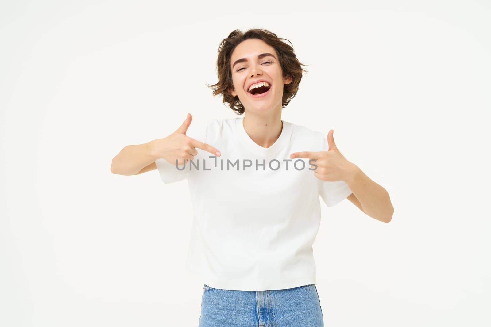 Image of brunette female model, pointing at herself and smiling, isolated against white studio background.