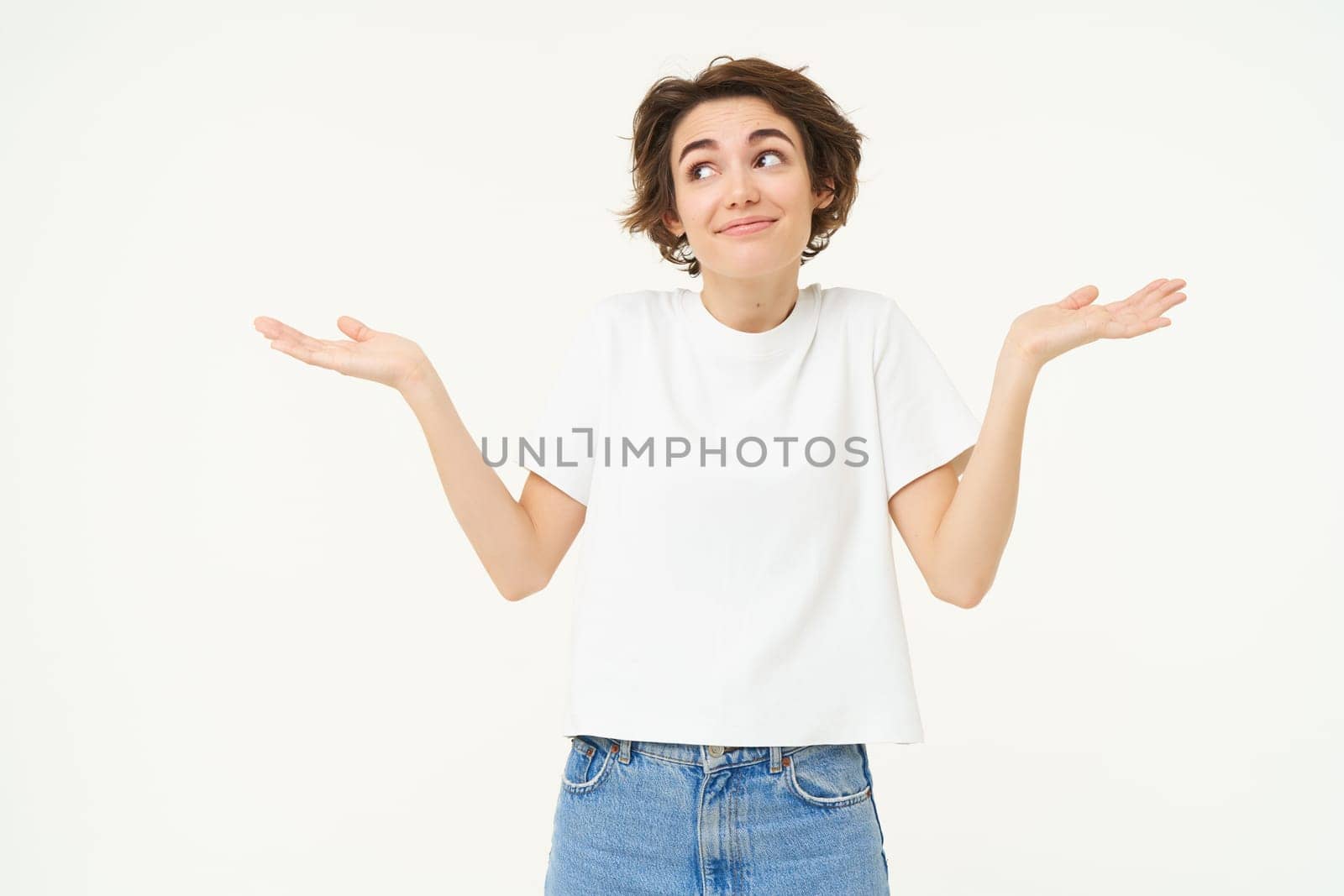 Portrait of girl with short hair, shrugs shoulders, looks unaware, clueless, doesnt know what to do, stands over white studio background.