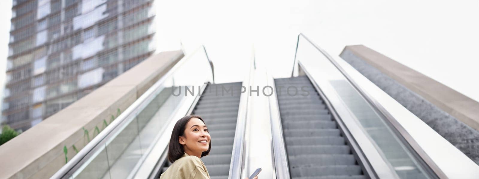 Young asian girl going up on an escalator, holding smartphone, smiling while walking in city. Copy space