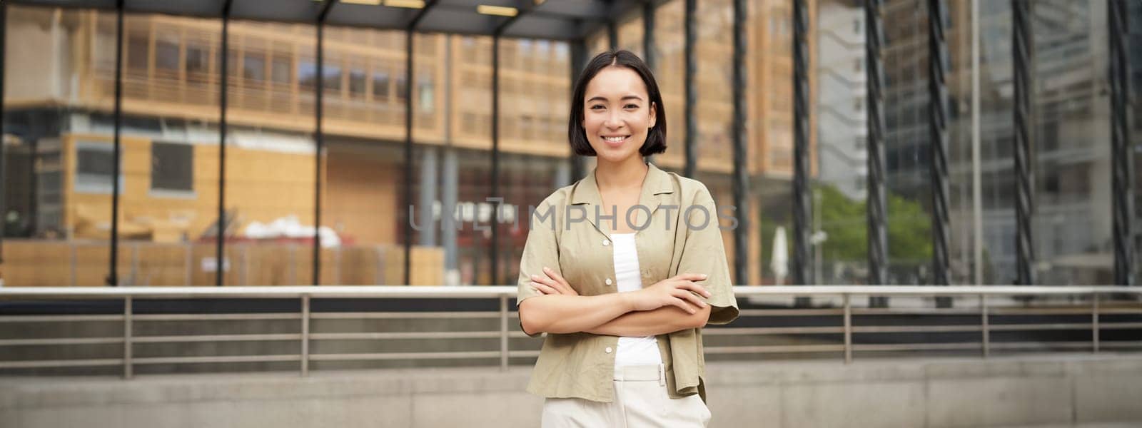 Portrait of beautiful young woman in casual clothes, smiling, posing outdoors on an empty street near glass building by Benzoix