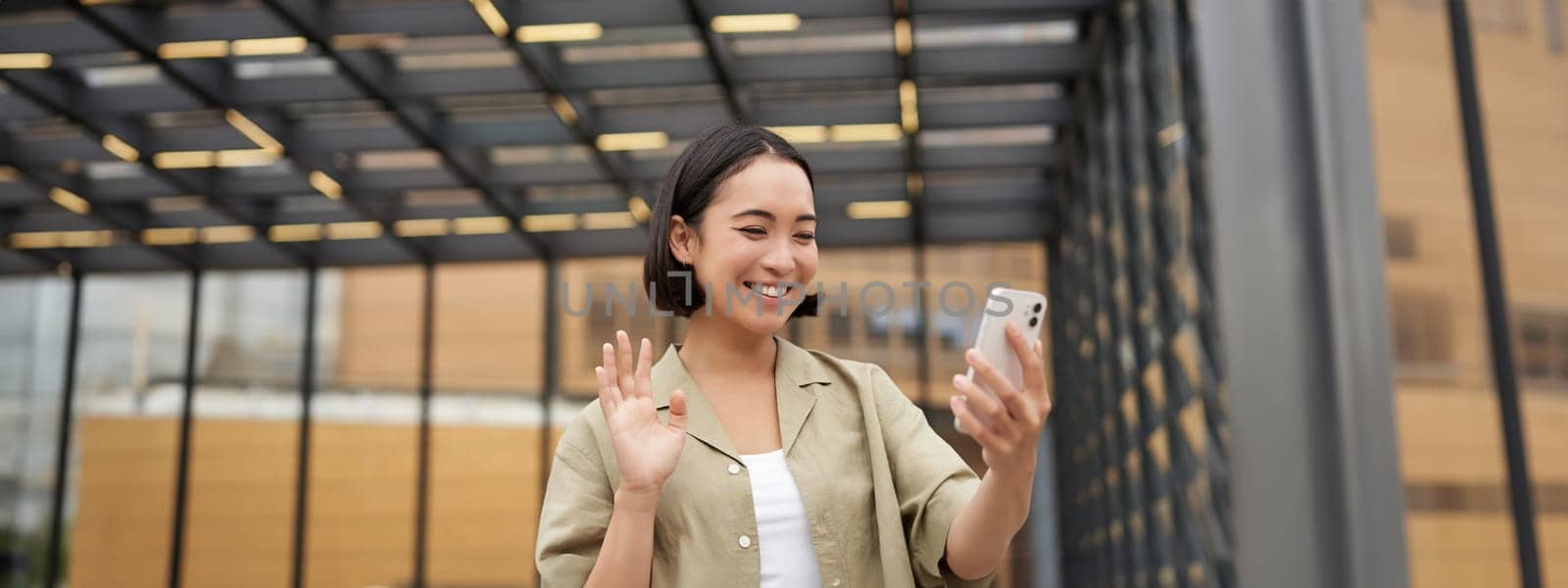 Happy asian girl waves at her smartphone camera, says hello to friend on video chat, calling someone, standing on street.