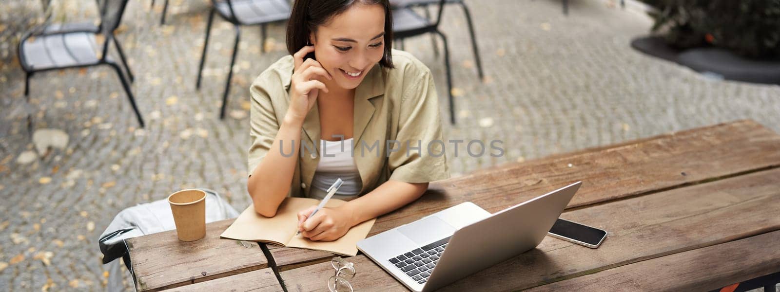 Portrait of young woman studying online, sitting with laptop, writing down, making notes and looking at computer screen, sitting in cafe outdoors.