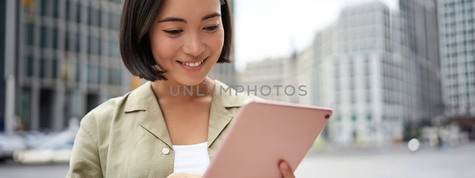 Portrait of asian woman reading, using tablet while standing on street, smiling while looking at screen by Benzoix
