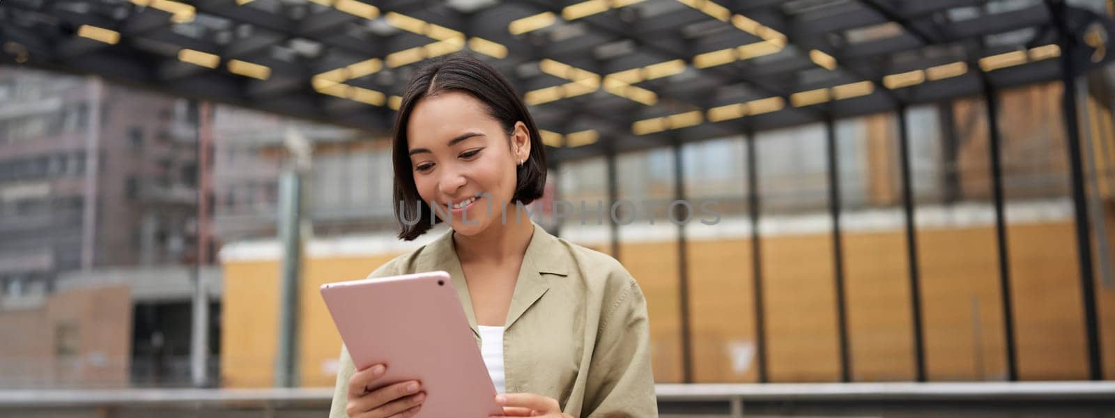 Portrait of beautiful asian girl using tablet, reading, looking at tablet while standing on street.