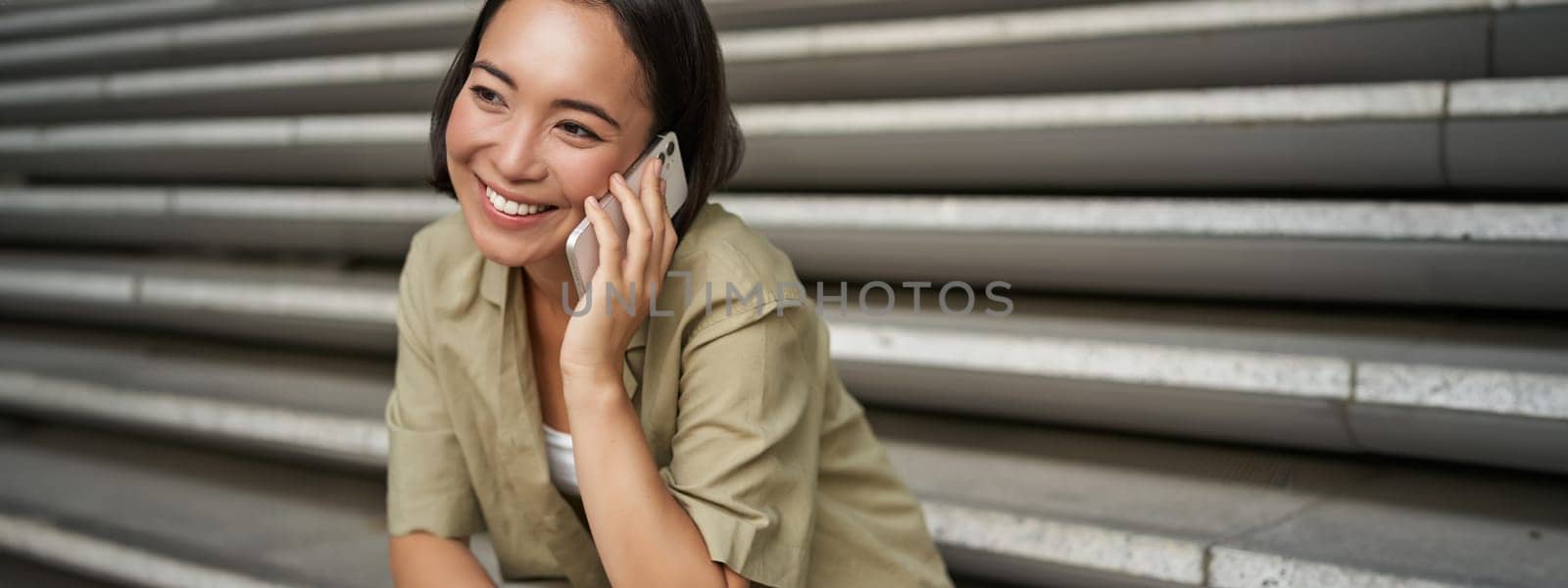 Close up portrait of smiling asian girl talks on mobile phone, sits outside on street stairs. Young woman calling friend on smartphone by Benzoix