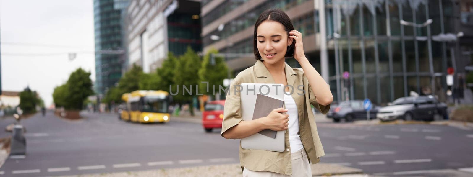 Portrait of beautiful asian woman standing on street with laptop and notebook, going to university or work, commute route.