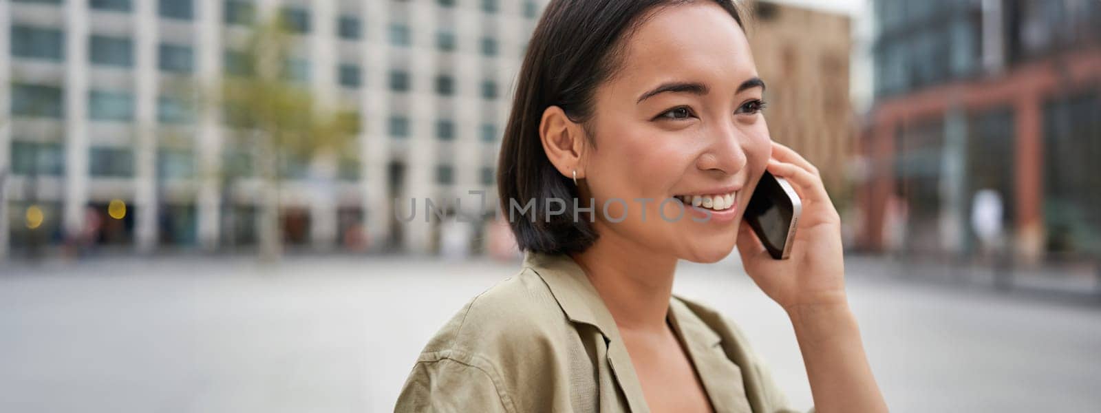 Modern young asian girl talks on mobile phone, uses telephone on city street. Woman smiling while calling someone on smartphone.