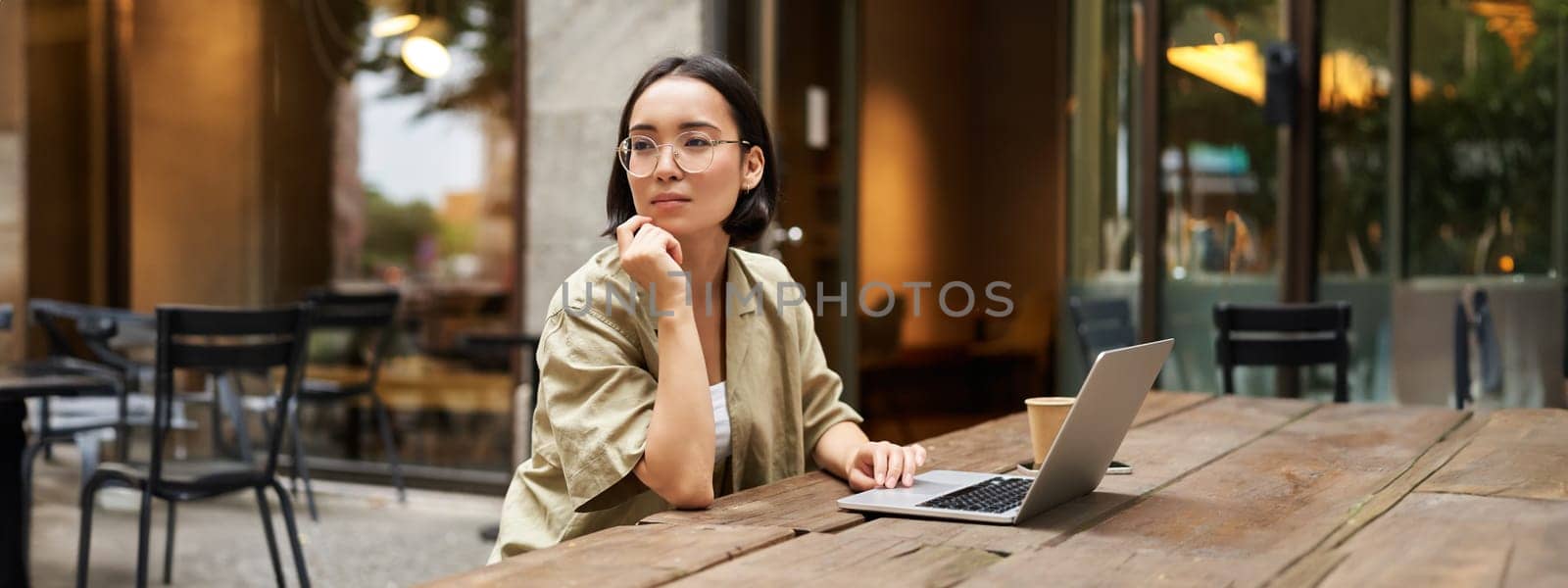 Working woman sitting with laptop in cafe and thinking. Asian girl in glasses works remotely, drinks coffee and looks thoughtful.