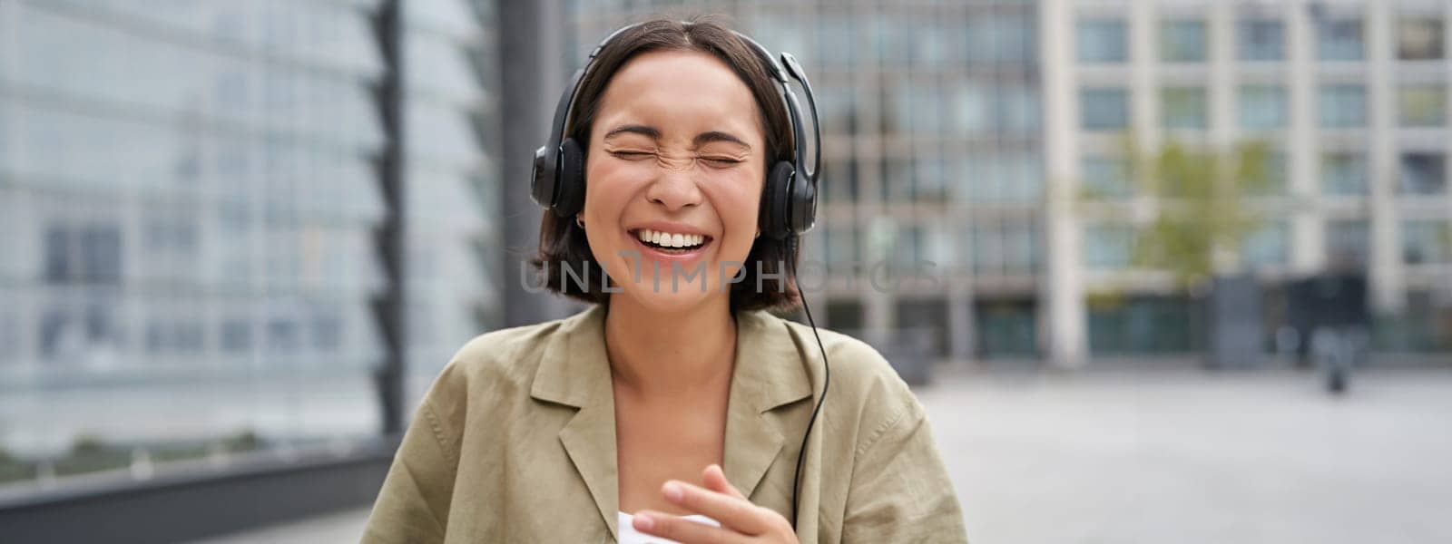 Carefree asian girl, laughing and smiling, wearing headphones and walking on street. Outdoor shot of young woman listening music and looking happy.
