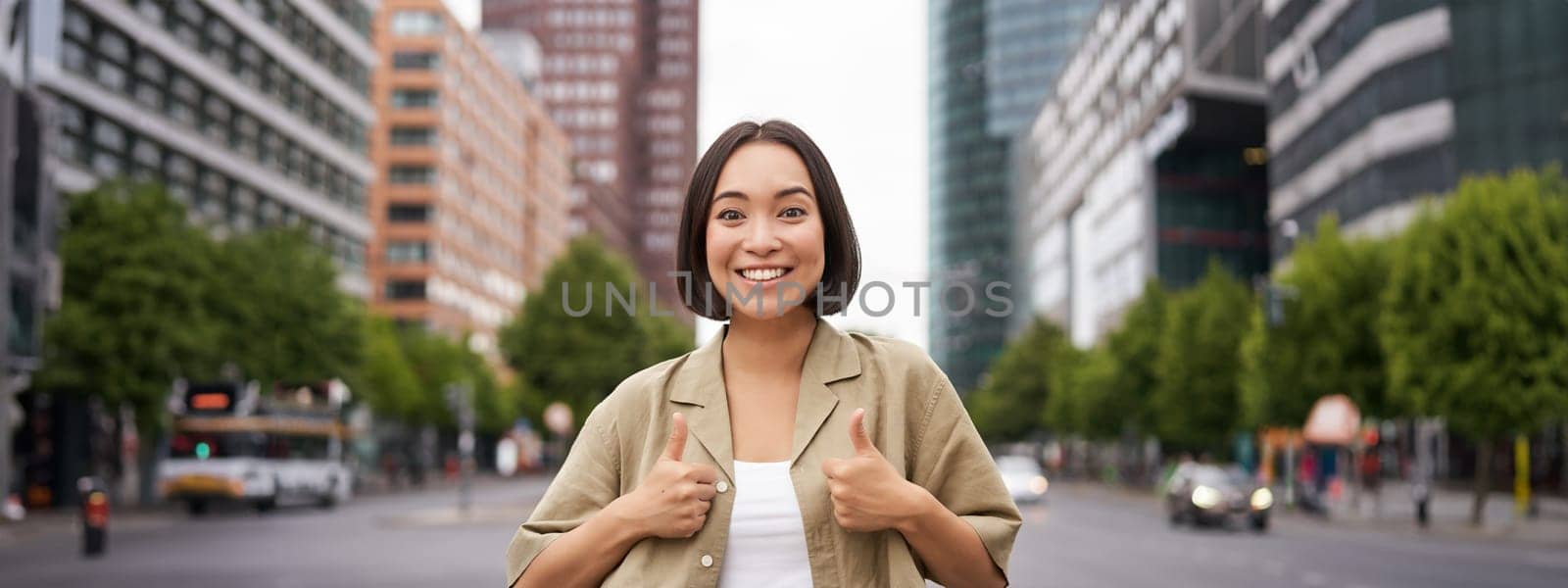 Enthusiastic city girl, shows thumbs up in approval, looking upbeat, say yes, approves and agrees, stands on street.