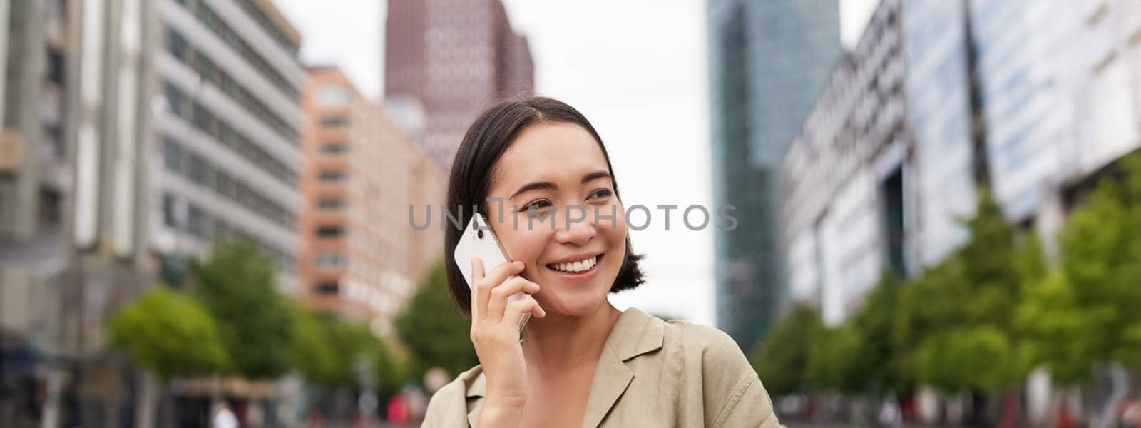 Portrait of smiling asian woman talking on mobile phone, walking on street near busy road and speaking to friend, laughing.