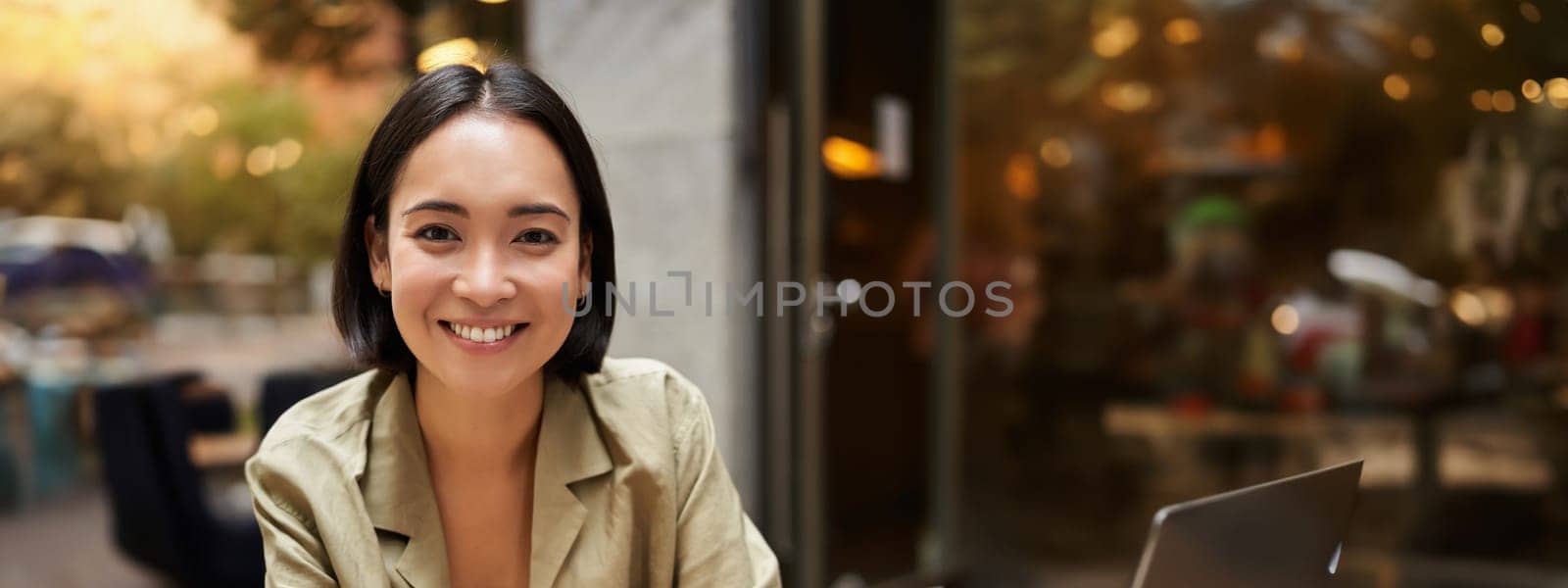 Vertical shot of young asian woman working on remote from outdoor cafe, sitting with laptop and smiling, studying.