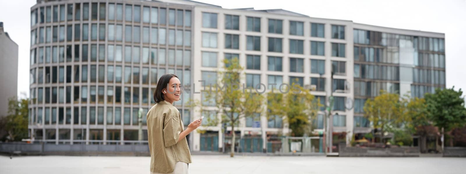 Silhouette of asian woman walking on street in wireless headphones, holding smartphone by Benzoix