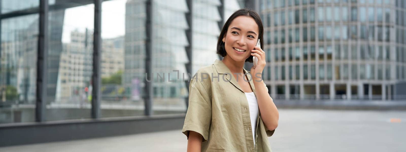 Portrait of asian girl smiling, talking on phone, making a call, standing on street near building and waiting for someone, answer telephone.