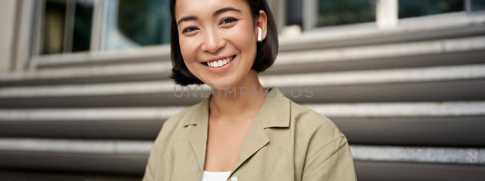 Portrait of smiling asian girl listens music, podast in wireless earphones, using headphones outdoors, sitting on street by Benzoix