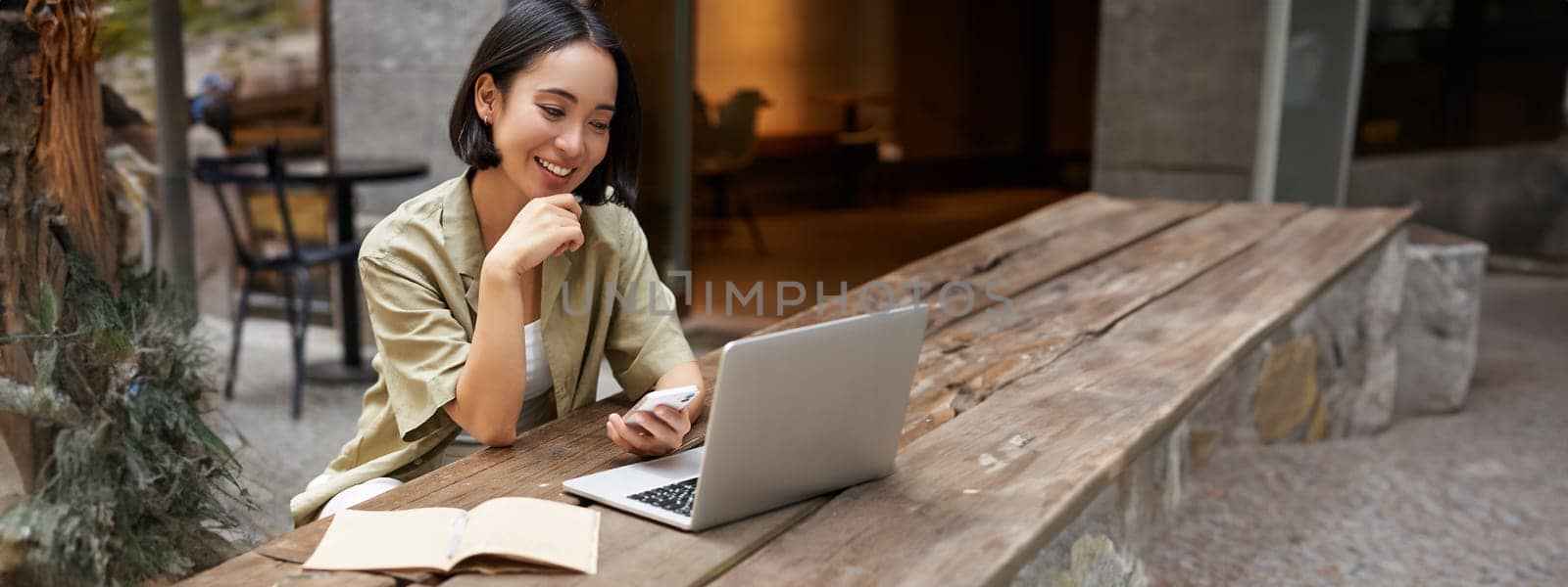 Portrait of stylish urban woman working on laptop from outdoor cafe, sitting with documents and computer, smiling by Benzoix