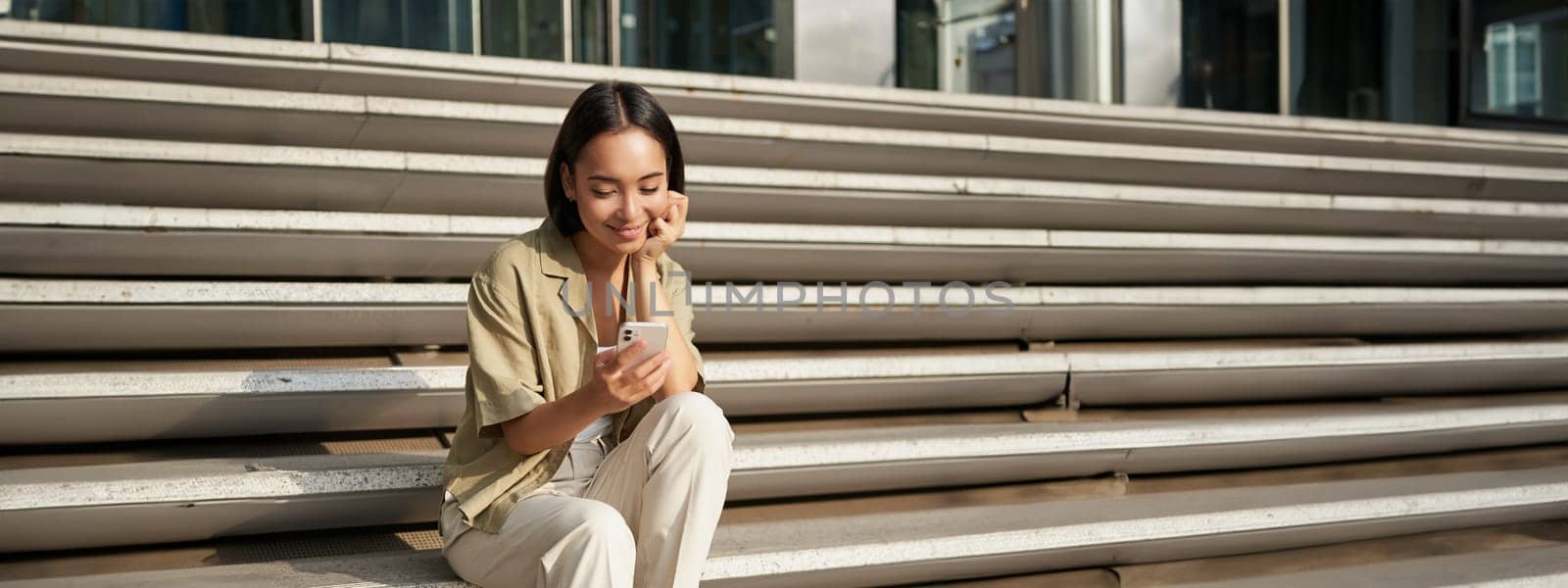Vertical shot of asian woman, student sits on stairs in city, looking at mobile phone screen and smiling, using smartphone app by Benzoix